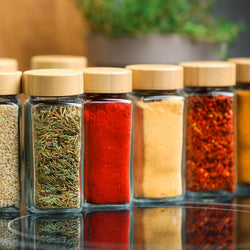 Verschiedene Bio-Gewürze in Glasgläsern mit Bambusdeckeln – Rosmarin, Paprika, Knoblauch und Chiliflocken in einer Reihe