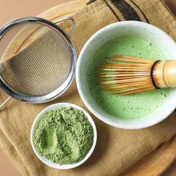 Matcha-Zubereitung mit Bambusbesen, Sieb und grünem Pulver in weißer Schale